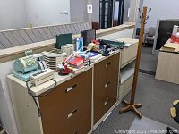 Overall lot view displaying numerous office items arranged on top of metal file cabinets. Wooden hall tree standing next to the cabinets. Visible items include keyboards, postage scale, office trays, calculators, binders, staplers and other small office tools.