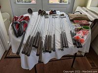 Wide view of 18 golf clubs, including putters and drivers, laid on a white tablecloth with three red and black head covers at the end.