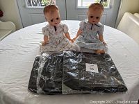 Two vintage twin dolls sitting on a white round table with three black bags stacked in original packaging in front of them.