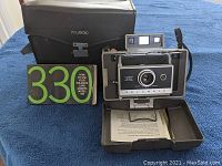 Front view of Polaroid Land Camera Model 330 alongside manual and carrying case