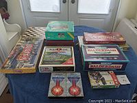 Overall assortment of board games and VHS tapes arranged on table including checker board, Scrabble, Hamilton and Balderdash.