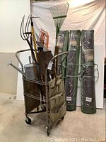 Front view of metal rolling cart loaded with various gardening tools including rakes, pitchforks, shovels, and watering can. Three bundles of rolled willow screening wrapped in plastic are placed behind.