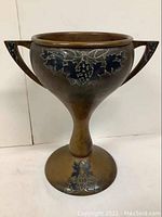 Full front view of copper urn showing decorative silver layover grapevine pattern and triangular handles.