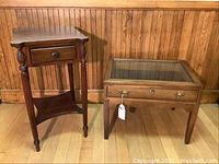 View of both wooden tables side by side, vitrine table on the right with glass display top and side table on the left with carved legs and drawer.