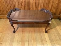 Full frontal view of the ornate wooden coffee table with glass top and Queen Anne legs on a wooden floor.