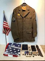 Olive drab US Army wool service jacket with staff sergeant chevrons, small US flag on stand, folded flags, Bluejackets' Manual, Silver Star case and assorted medals, ribbons and insignia displayed