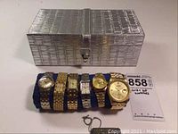 Seven gold plate and two-tone wristwatches displayed on navy blue watch roll with textured silver jewelry box in back.