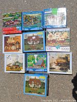 Twelve boxed 1000-piece jigsaw puzzles displayed on concrete, mostly Hometown Collection and Charles Wysocki puzzles, visible box artwork shows various Americana village and home scenes.