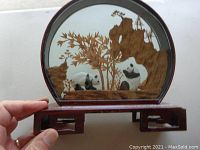 Photo showing the 3D cork art piece with panda bears in a scenic setting with a wooden base, held by hand for size reference.