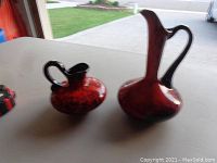 Two red and black Blue Mountain Pottery pitchers shown together on a white surface with outdoor background.