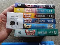 Hand holding five Hawaii Five-0 DVD box sets stacked showing spines of seasons 1 to 5.