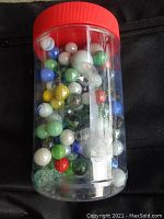 Jar filled with assorted glass marbles in multiple colors and styles with red lid on top.