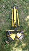 Two yellow metal worklamps on adjustable poles lying on grass. One worklamp missing bulb. Attached cords visible.