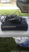 Photo showing black Xbox 360 console with cables on top, placed on a table outdoors, with a plastic bag underneath.
