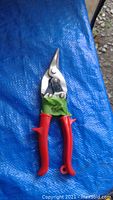 Close-up photo of a single pair of tin snips on a blue tarp. The snips have silver metal blades and red handles with green tape wrapped around the joint area.