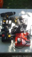 Multiple cordless drills, chargers and batteries laid out on table outdoor, showing condition and mismatched parts