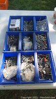 Photo showing three blue trays each divided into compartments filled with various sizes and types of screws, bolts, washers, nuts and other small hardware parts in new condition.