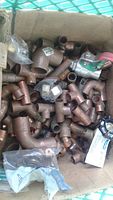 Box filled with various copper plumbing fittings including elbows, tees, reducers, and a few valves with red handles