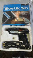 Black Bostik 260 electric glue gun with power cord in molded styrofoam tray, placed inside the original blue and white Bostik 260 box with text in multiple languages.