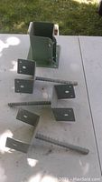 Photo showing 3 large green L-shaped fence brackets with concrete inserts and 1 small metal bolt down bracket laid out on a white table against grass background.