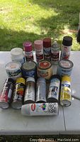 Photo showing multiple spray paint cans and paint cans on white surface outdoors.