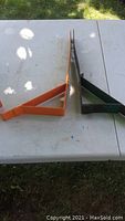 Two metal roof jacks on a white table, one orange and one green, viewed from the side showing their L-shaped design.