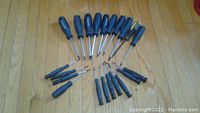 Photo showing approximately 21 torque screwdrivers arranged in a semi-circle on a wooden floor, showing black handles and metal shafts with various head types and sizes.