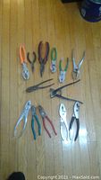 Photo showing 10 various pliers arranged on a wooden floor, with visible different handle colors and types of pliers.