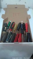 Photo showing a box of 12 assorted used screwdrivers with different handle colors including black, red, green, and transparent yellow, displaying flathead and Phillips heads.