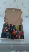 Photo showing a cardboard box containing about 19 assorted screwdrivers of varying sizes and handle colors.