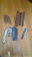 Photo showing a collection of metal punches, flat cold chisels, two utility knives, a scraper, a pry tool, and an Allen wrench laid out on a wooden floor.