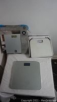 Three floor scales displayed on a white surface: one in box, one white analog scale, one gray digital scale