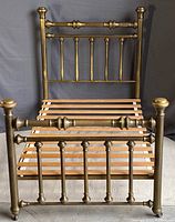Front view of antique brass bed frame showing headboard and footboard with vertical spindles and horizontal upper rails, wooden slats visible.
