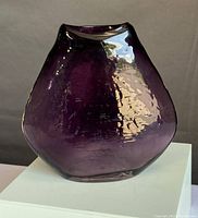Front view of a violet hand-blown art glass vase with an uneven organic shape on a white pedestal against a dark background.