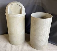 Two beige/gray Rubbermaid garbage bins side by side, one complete with dome-shaped lid, liner removed and set inside, and one empty base barrel