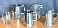 Five assorted beer steins displayed on a wooden surface. Visible are pewter steins with hinged lids, glass bottoms, and one etched glass stein.