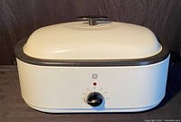 Front view of white GE roaster oven with temperature control dial and red indicator light, black trim, and lid knob visible.