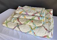 Folded quilt with three pillow shams stacked on top, showing colorful lattice ribbon design on cream background.
