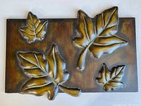 Front view of rectangular metal artwork featuring four embossed, golden-bronze leaves on a dark background.