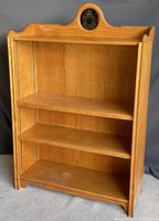 Front view showing oak bookshelf with two adjustable shelves and decorative crest on top center, wood grain and light wear visible.