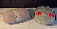 Both foot massagers side by side on a white surface, one beige with fabric covering and blue power button, the other grey with red heat lights.