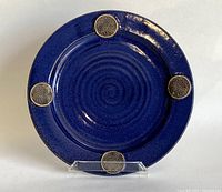 Front view of blue stoneware pottery plate with four Celtic knot medallions and spiral center texture, on clear stand.