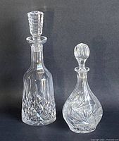Two cut crystal decanters shown side by side, one pinwheel pattern and ball stopper, one tall diamond cut with geometric stopper.