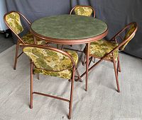Four vinyl-upholstered folding chairs with floral pattern arranged around round card table with green laminate top and copper-toned metal frame.