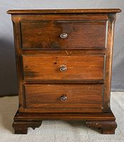 Front view of the wooden bedside table showing three drawers and the metal drawer pulls, surface shows some wear.