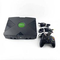 Front view of the black original Xbox console, showing controller ports and the green logo on top, with included AV and power cables and a wired controller next to it.