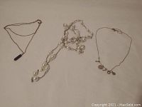 Three necklaces laid out on flat surface: pearl strand, delicate chain with I Love You charms, and thin chain with dark pendant.