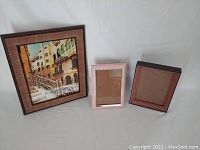 Photo showing three picture frames on a white background. Left frame is large brown wooden frame with street scene print. Middle frame is small pink metal photo-size frame. Right frame is small dark brown wooden frame with reddish inner border.
