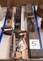 Three vintage wood block planes, a vintage sad iron, and a box with assorted plane parts displayed on a table.