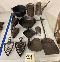 Wide view of lot on white table showing various cast iron and tin items including trivets, bail pot, flat irons, skillet, strainers and shovel